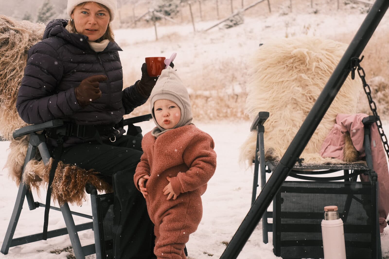 En mor med kaffekopp og et barn i ulldress i snøen